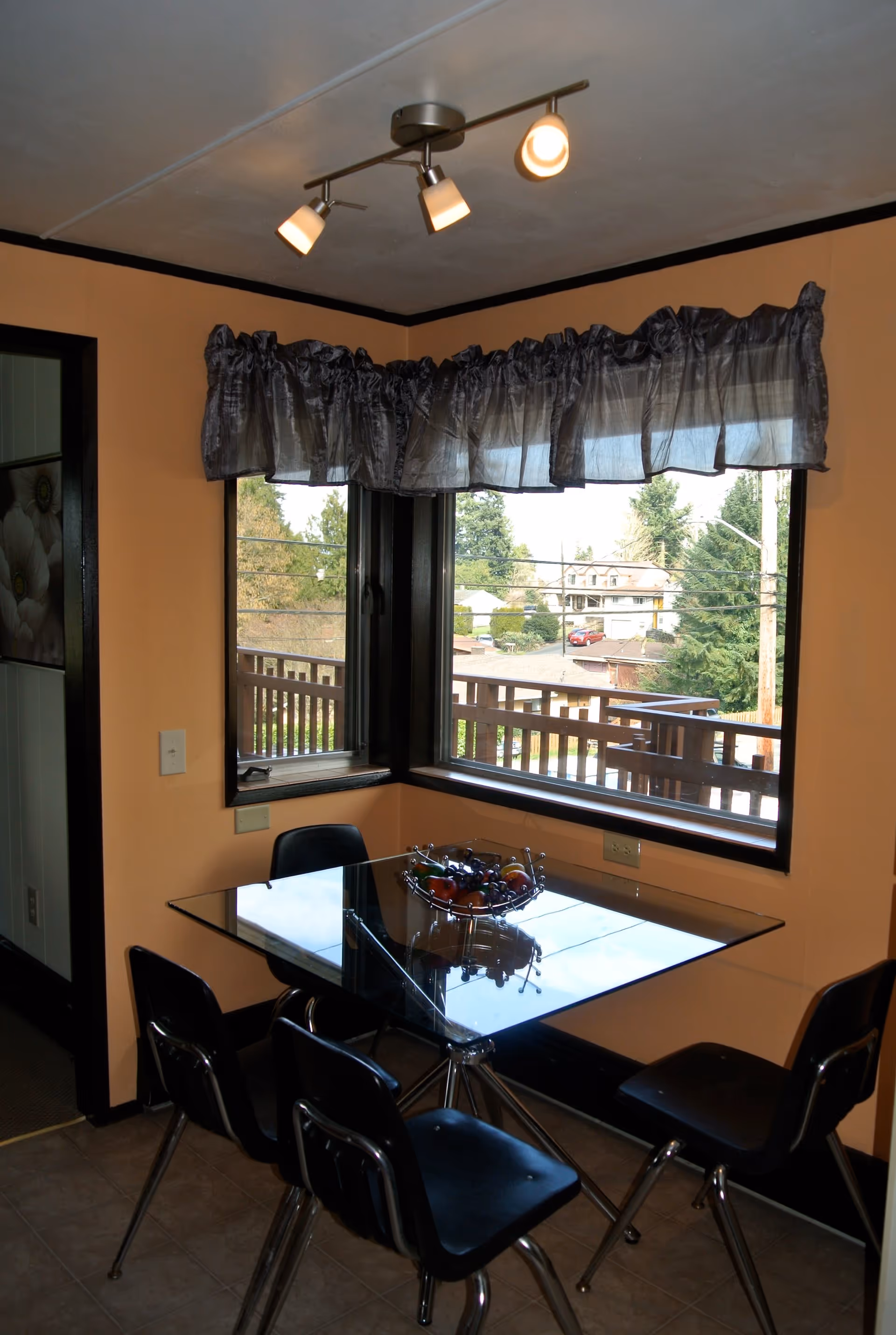 A small dining area with a glass square table and four black chairs. A decorative fruit bowl is placed on the table. The walls are painted peach with black trim around the windows and door. Two large windows with sheer dark curtains provide a view of trees and houses outside. A ceiling light fixture with three adjustable spotlights is mounted above the table.