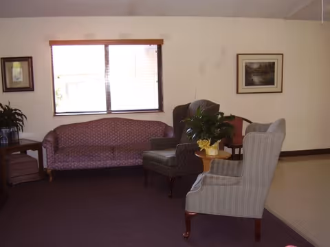 A small sitting area in a senior care facility with a patterned sofa, two upholstered armchairs, a wooden side table with a potted plant, and framed artwork on the walls. A window with blinds lets in natural light.