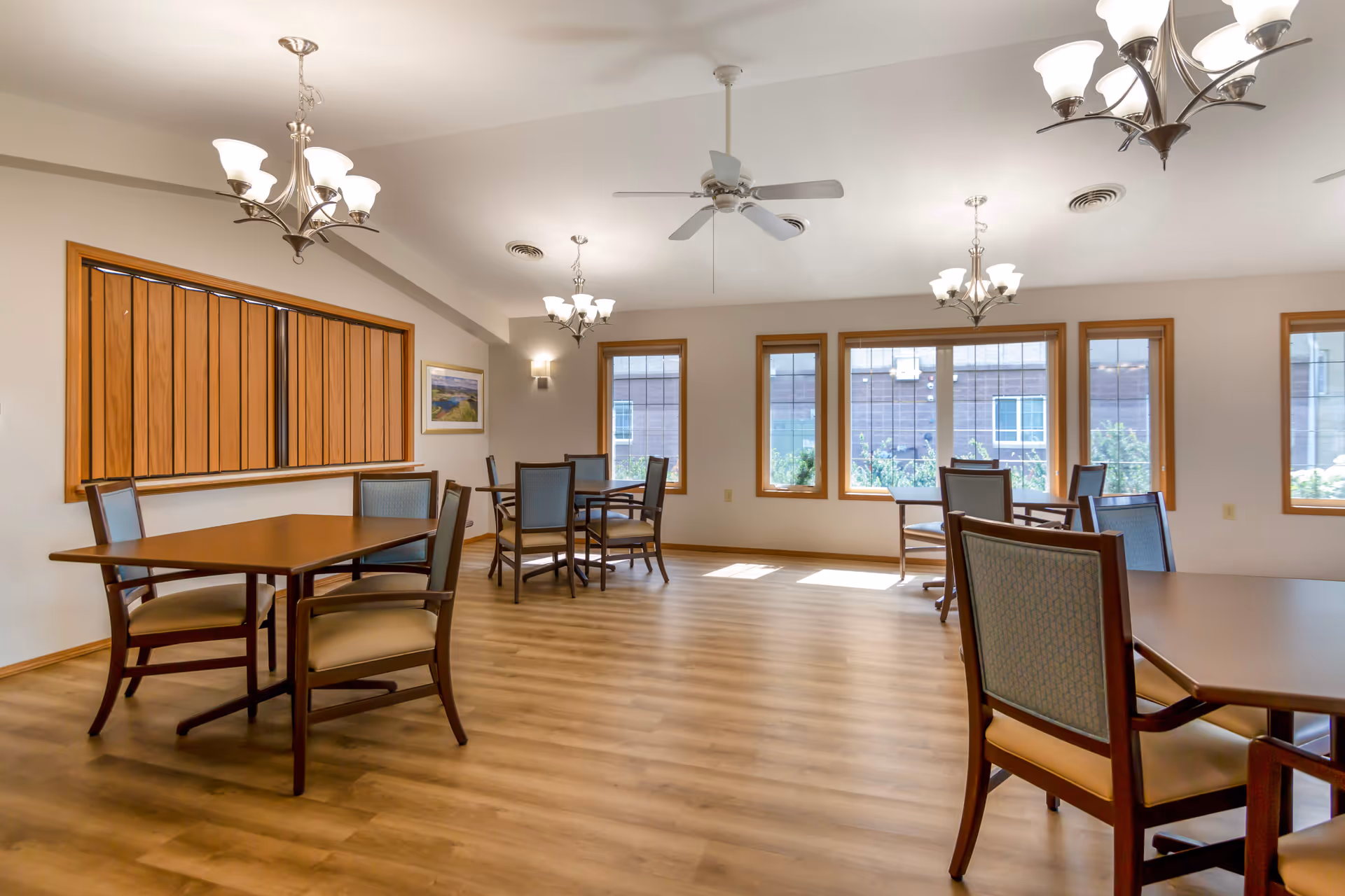 A bright dining room with several wooden tables and chairs arranged on a wood floor. The room has large windows with wooden frames letting in natural light, and multiple ceiling light fixtures and a ceiling fan are visible. There is a framed picture on the wall and a wooden panel feature on one side.