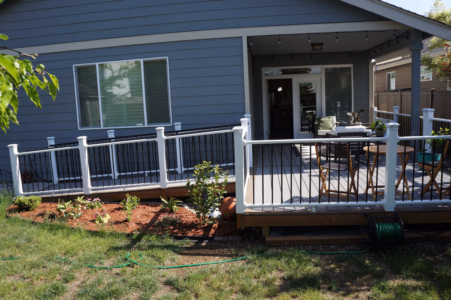 A backyard patio area of a house with a wooden deck and white railing with black balusters. The deck has outdoor furniture including chairs and a small table. There is a garden bed with plants and mulch in front of the deck, and a green garden hose is coiled on a holder beneath the deck. The house exterior is gray with a large window and a sliding glass door leading to the patio.