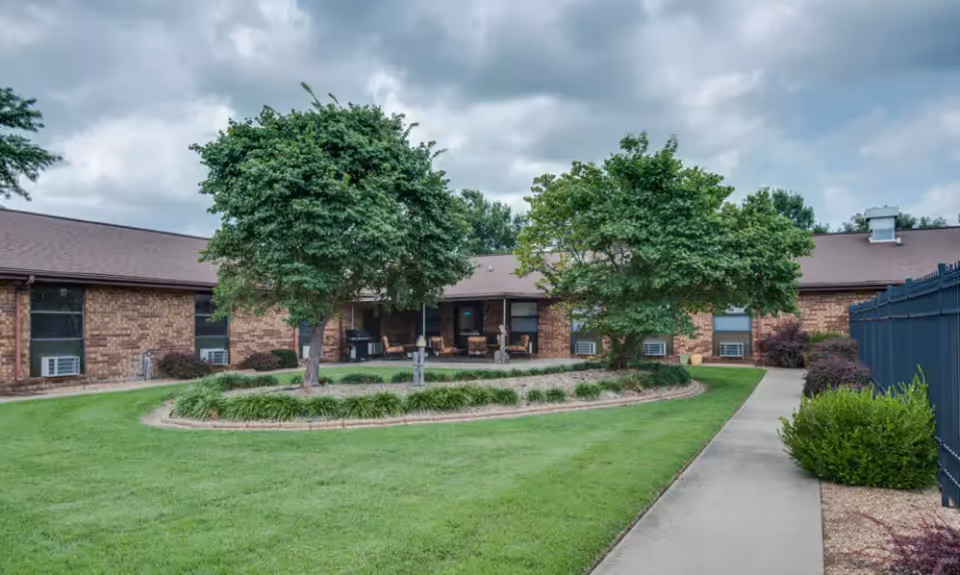 Outdoor courtyard area of a nursing center with a well-maintained lawn, two large trees, a circular garden bed, a paved walkway, and a brick building in the background under a cloudy sky.
