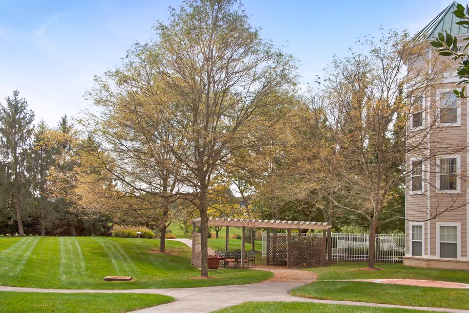 Outdoor area of a senior living facility with a well-maintained lawn, several trees with sparse leaves, a paved walkway, and a wooden pergola with seating underneath. Part of a multi-story building with beige siding and white-trimmed windows is visible on the right side.