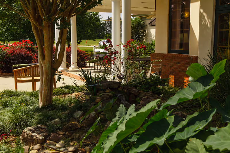 Outdoor patio area at an assisted living facility featuring a small garden with large green leaves, a tree, rocks, and a water feature. There are wooden benches and metal tables with chairs under a covered porch supported by white columns. Red flowering bushes and a grassy lawn are visible in the background.