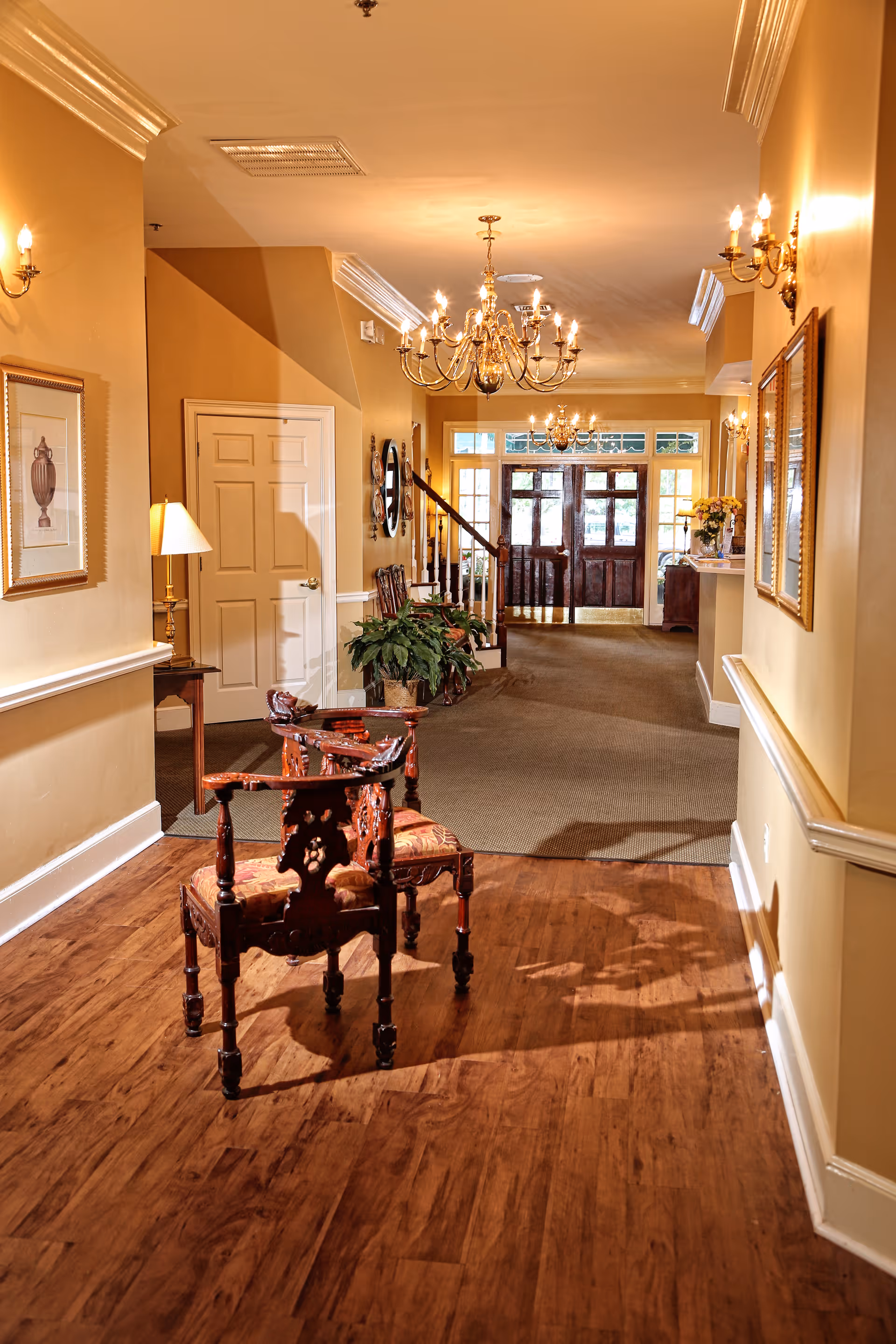 Hallway interior of a senior living facility with wooden chairs, chandeliers, framed art, and front entrance doors.