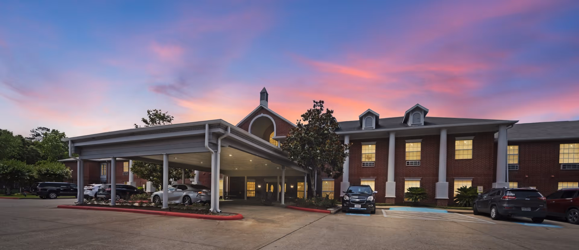 Exterior view of Collier Park facility at sunset with a covered entrance, several parked cars, and a two-story brick building with lit windows and white columns.