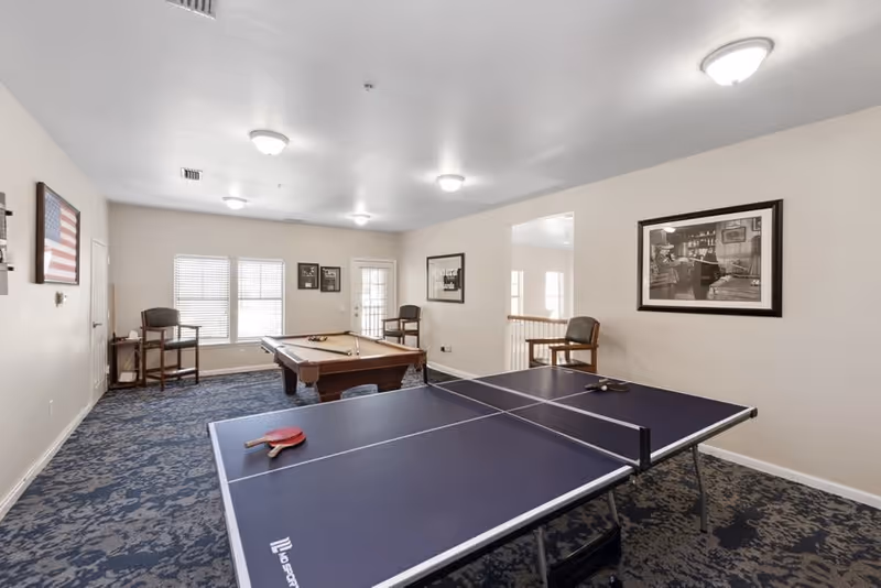A recreational room with a ping pong table in the foreground and a pool table in the background. The room has patterned blue carpet, beige walls, and several framed pictures on the walls. There are chairs placed around the room and windows letting in natural light.