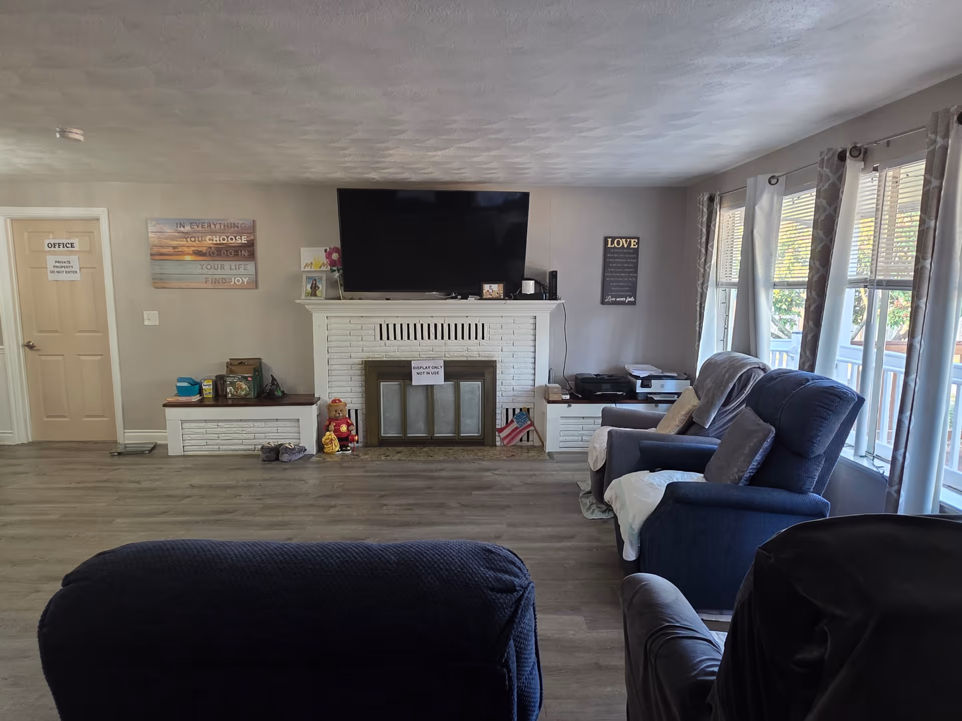 Spacious living room with a TV above a white brick fireplace, recliner chairs, and large windows along the right wall.