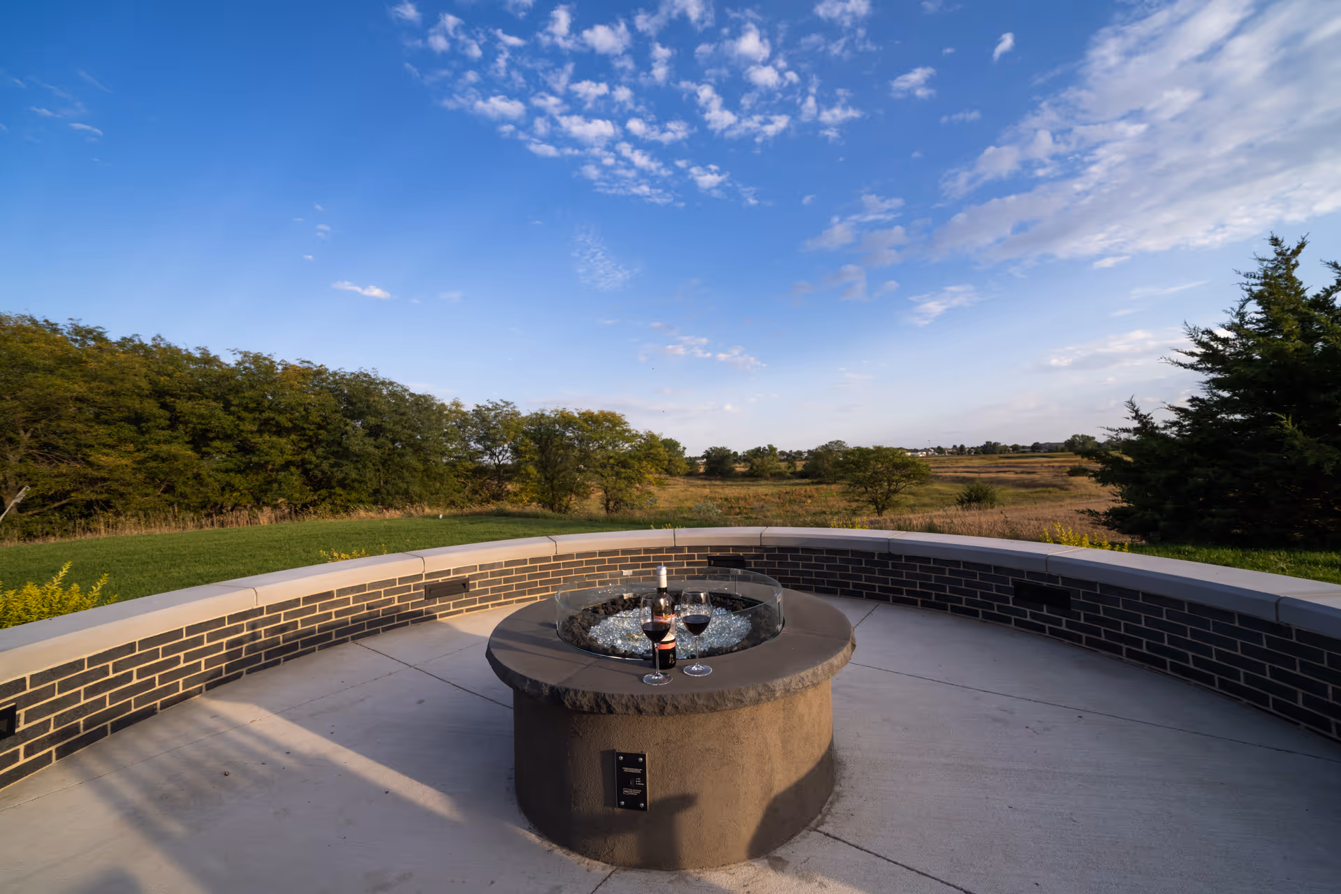 Outdoor patio area with a circular fire pit table in the center, holding a bottle of wine and two glasses filled with red wine. The patio is surrounded by a curved brick wall with a scenic view of trees, grass, and a partly cloudy blue sky.