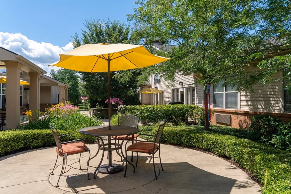 Outdoor patio area with a round table and three chairs with red cushions under a large yellow umbrella. The patio is surrounded by green bushes, flowering plants, and trees. Residential buildings with beige siding and white trim are visible in the background under a blue sky with some clouds.