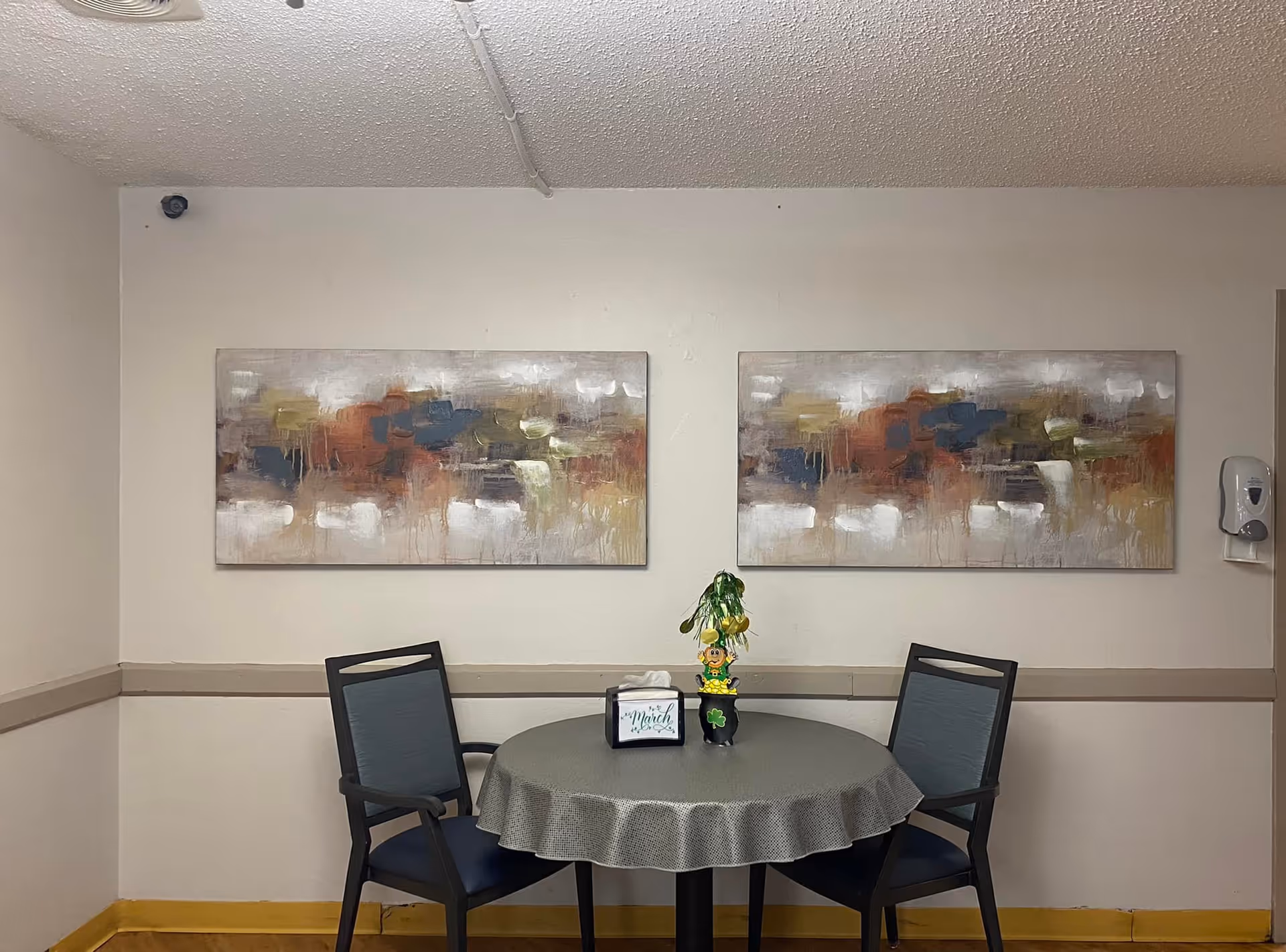 A small round table covered with a gray tablecloth is set against a beige wall. Two black chairs with blue cushions are placed on either side of the table. On the table, there is a tissue box and a small decorative plant with a leprechaun figure. Above the table, two abstract paintings with a mix of muted colors hang side by side. A wall-mounted hand sanitizer dispenser is visible on the right side of the wall.