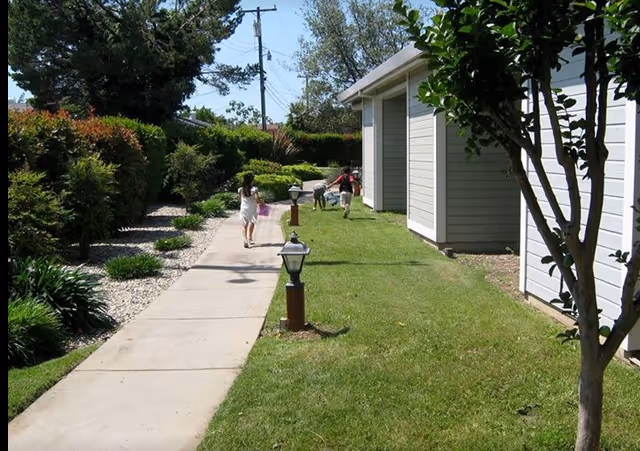 Children walking along a paved walkway beside white single-story buildings and landscaped lawns.