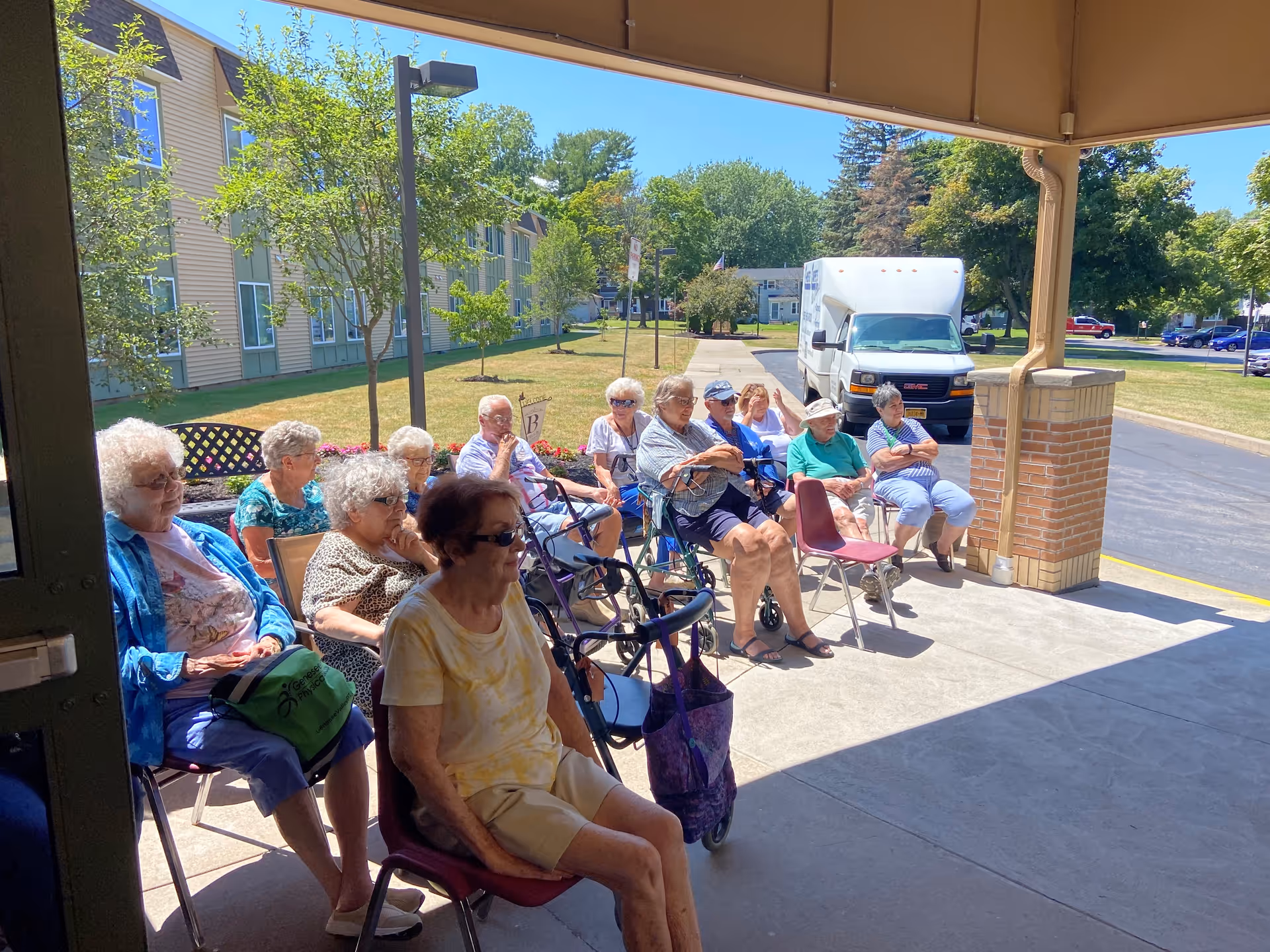 A group of elderly people sitting on chairs outside under a covered area near a building. They are seated in a row facing the open space, with some using walkers. Behind them is a parking lot with a white van and trees in the background on a sunny day.