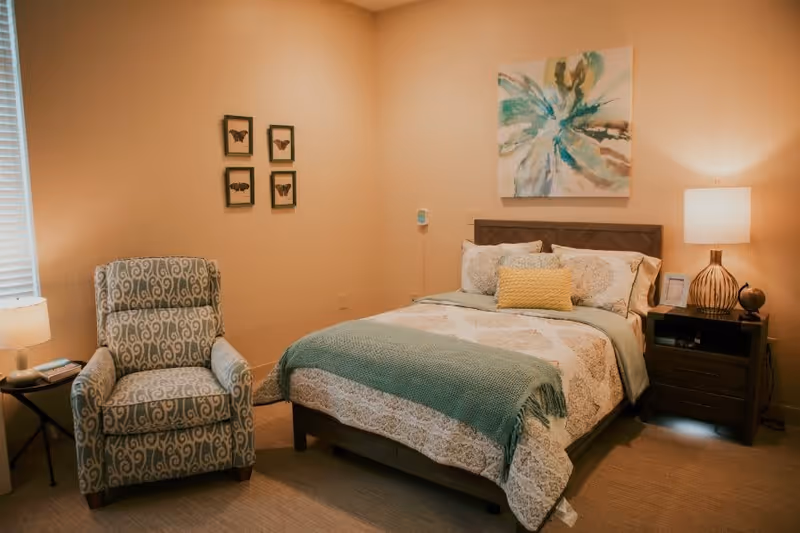 A cozy bedroom with a bed featuring patterned bedding and a green throw blanket. There is a wooden nightstand with a lamp and decorative items on the right side of the bed. To the left, there is a patterned armchair next to a small round table with a lamp and a phone. The walls are beige with framed butterfly artwork and a large abstract painting above the bed.