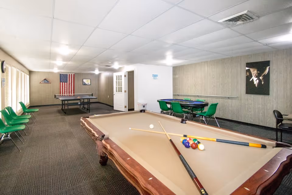 A recreational room with a pool table in the foreground, a ping pong table in the background, and a card table with green chairs along the right wall. The room has beige walls, a carpeted floor, and several green chairs lined up along the left wall under windows. An American flag and a folded flag display case are mounted on the far wall.