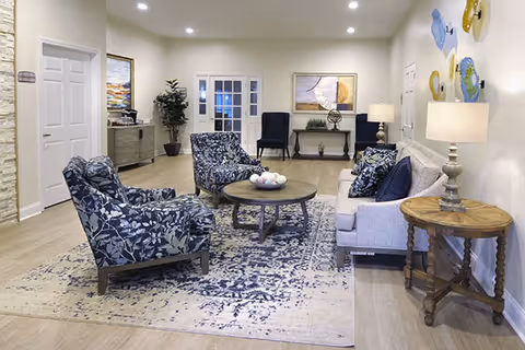 A well-lit living room with a light gray sofa, two blue and white patterned armchairs, a round wooden coffee table with decorative items, and a wooden side table with a lamp. The room features light wood flooring, a large patterned area rug, wall art, and recessed ceiling lights. There are two dark blue chairs and a console table with decor against the far wall, along with a potted plant and a door with glass panels.