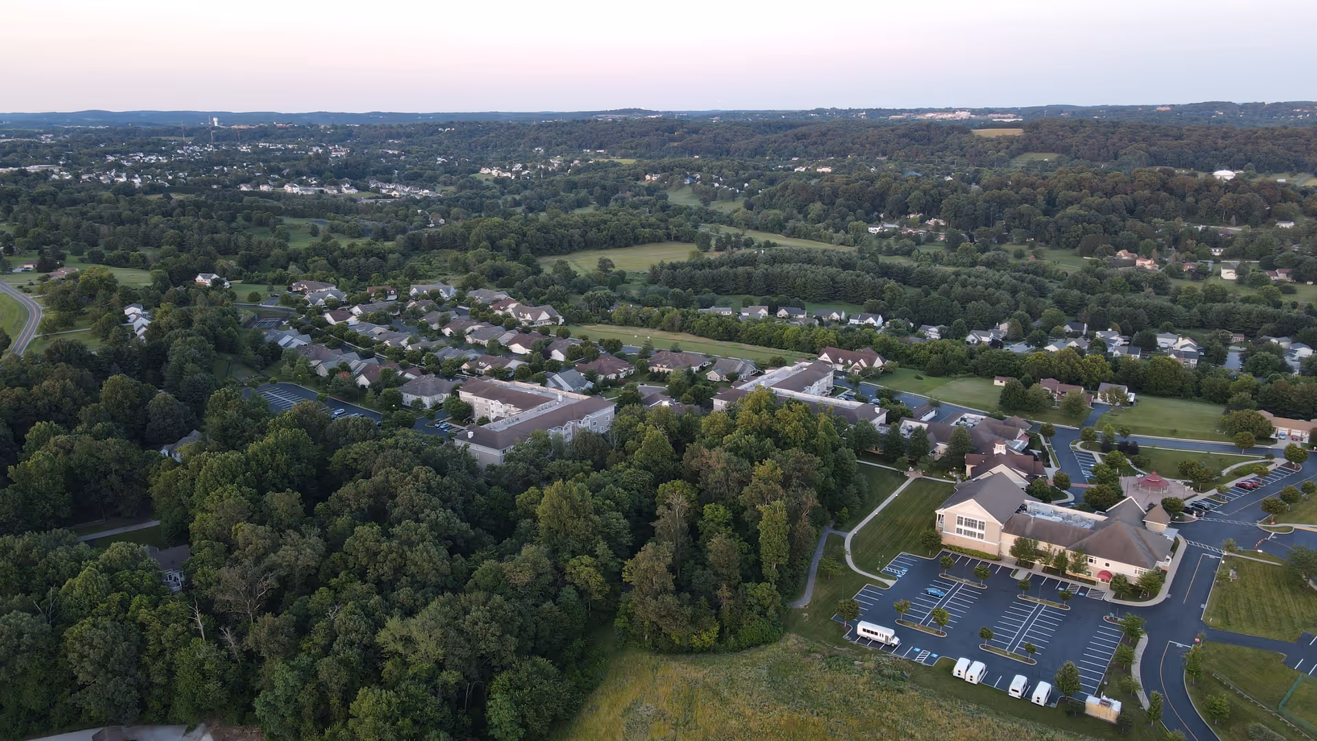 Aerial view of Carroll Lutheran Village showing multiple residential buildings surrounded by trees and greenery, with parking lots and roads visible. The landscape extends to distant hills under a clear sky.