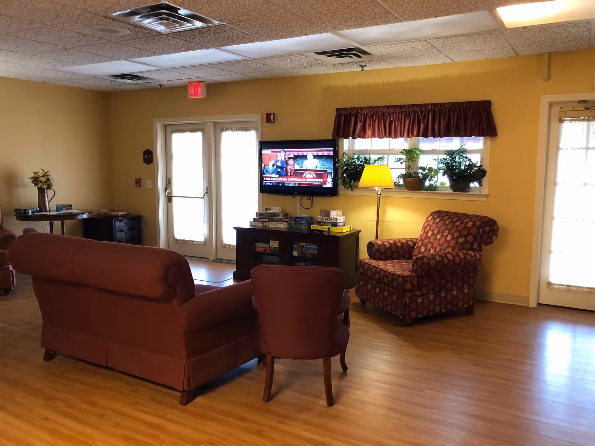 A cozy living room area with a maroon sofa, a matching armchair with a floral pattern, and a smaller chair. There is a wooden cabinet with board games on top and a flat-screen TV mounted on the wall showing a news program. A floor lamp is next to the armchair, and a window with a maroon valance has several potted plants on the windowsill. The room has wooden flooring and yellow walls, with double glass doors letting in natural light.