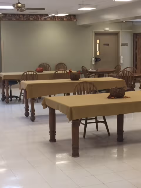 A dining area with several wooden tables covered with beige tablecloths and wooden chairs around them. Small decorative pumpkins are placed on the tables. The room has a white tiled floor, a ceiling fan, and fluorescent lighting. A door and a hand sanitizer dispenser are visible in the background.