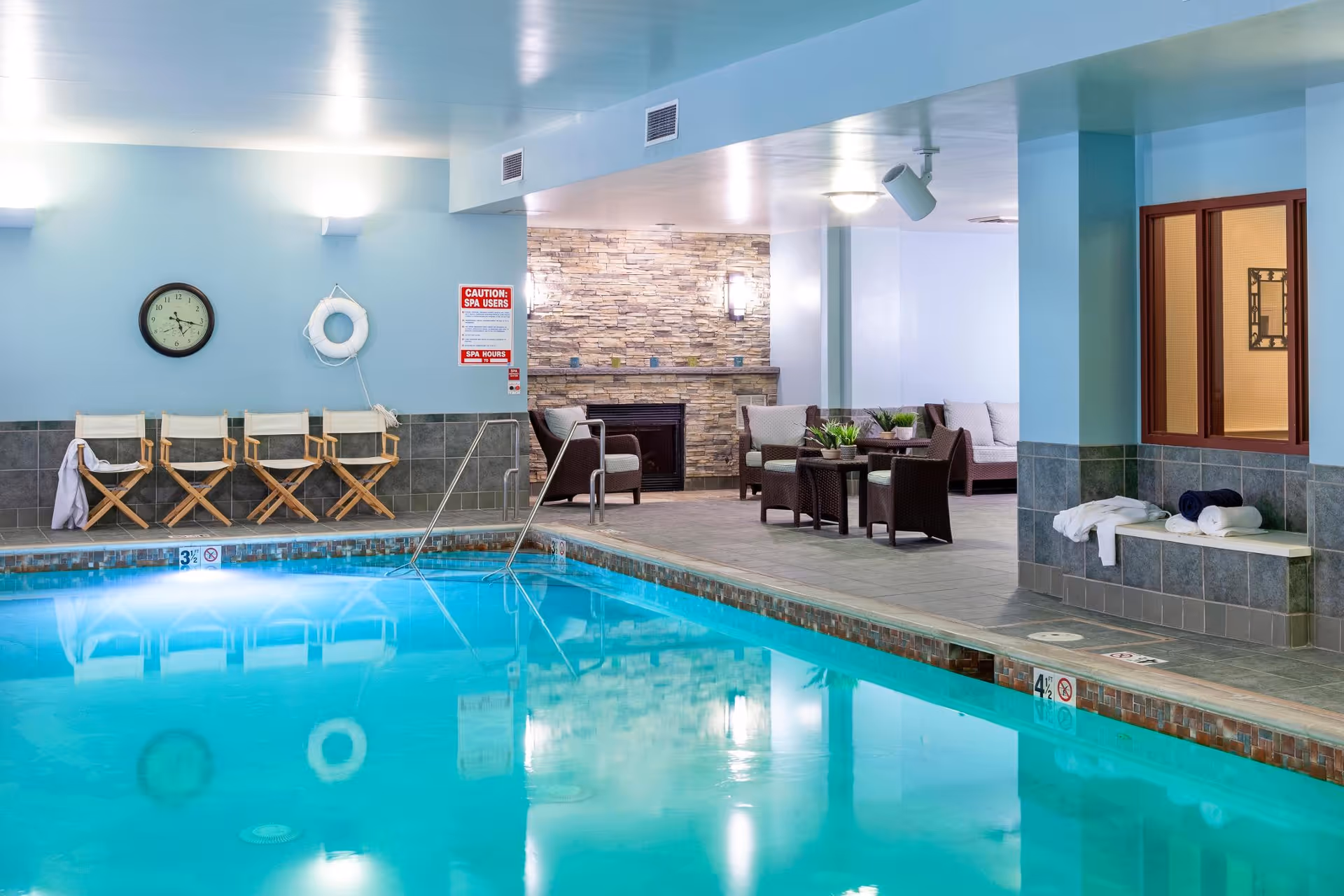 Indoor swimming pool area with clear blue water, surrounded by tiled floor. On one side, there are four wooden folding chairs lined up against a light blue wall with a clock and a life preserver hanging. On the other side, there is a seating area with wicker chairs and a sofa around a small table, with a stone fireplace in the background. Towels are placed on a bench near a window.