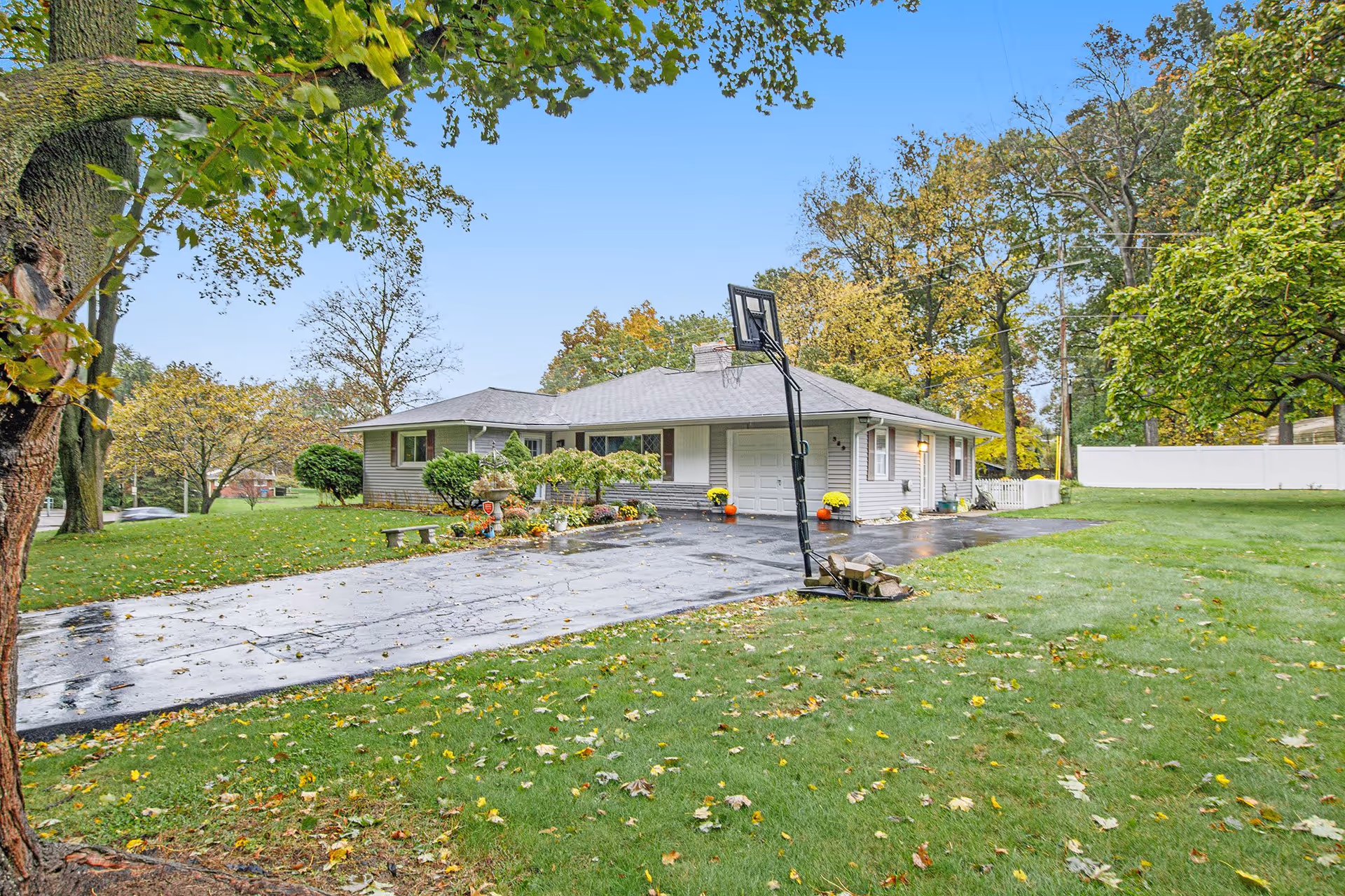 A single-story house with a gray roof and light-colored siding surrounded by green grass and trees with autumn leaves. There is a paved driveway with a portable basketball hoop and some flower pots near the entrance. The sky is clear and blue.
