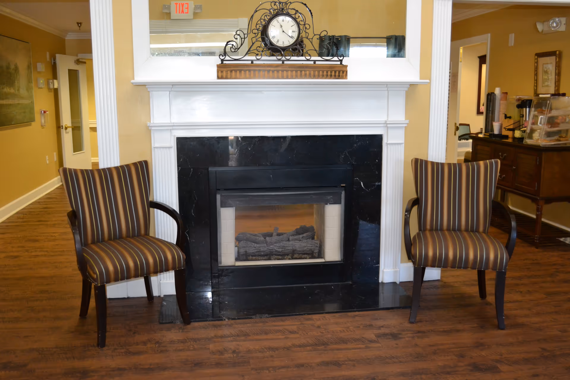 A cozy interior space featuring a black marble fireplace with a white mantel. On the mantel sits a decorative clock with an ornate metal frame. Two striped armchairs with wooden arms are positioned on either side of the fireplace. The room has wooden flooring and beige walls, with a hallway visible to the left and a small table with coffee supplies to the right.