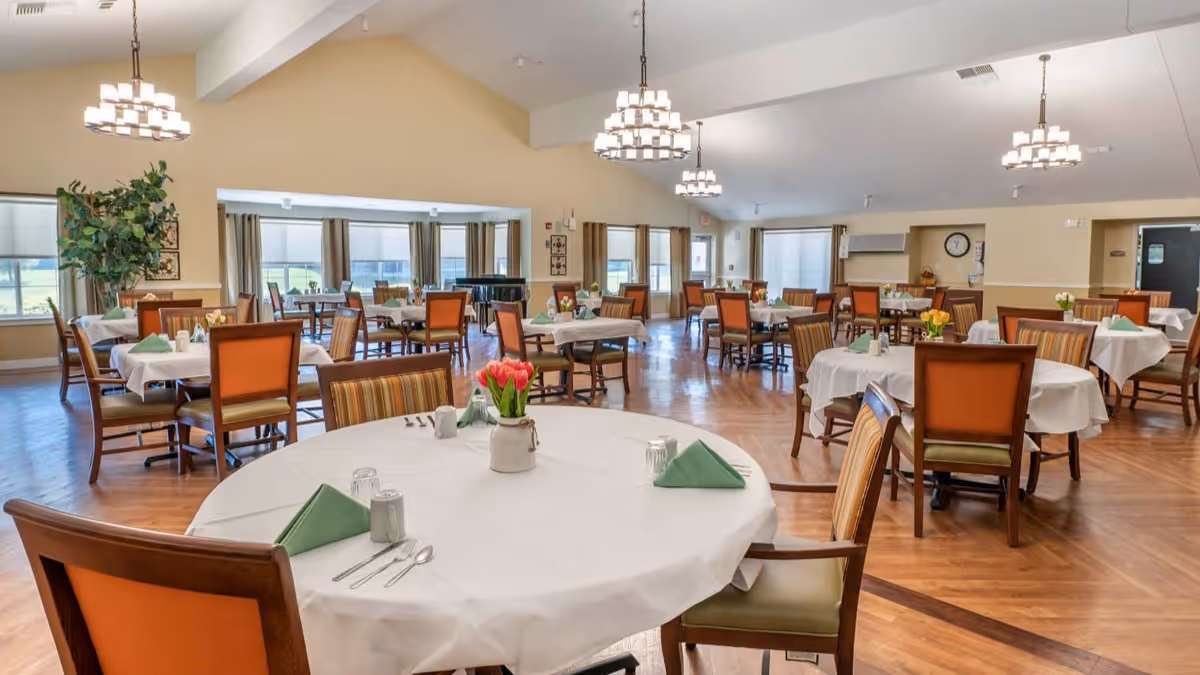 Spacious dining room with multiple round and square tables covered with white tablecloths, each set with green folded napkins, glasses, and silverware. Chairs with orange and striped upholstery surround the tables. The room has wooden flooring, large windows with beige curtains, and modern chandeliers hanging from the ceiling. There is a large plant in one corner and a piano near the windows.