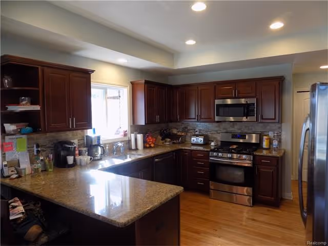 A modern kitchen with dark wooden cabinets, granite countertops, stainless steel appliances including a refrigerator, microwave, and gas stove. The kitchen has a window above the sink letting in natural light, and various kitchen items such as a coffee maker, paper towels, and fruit bowl are visible on the countertops.
