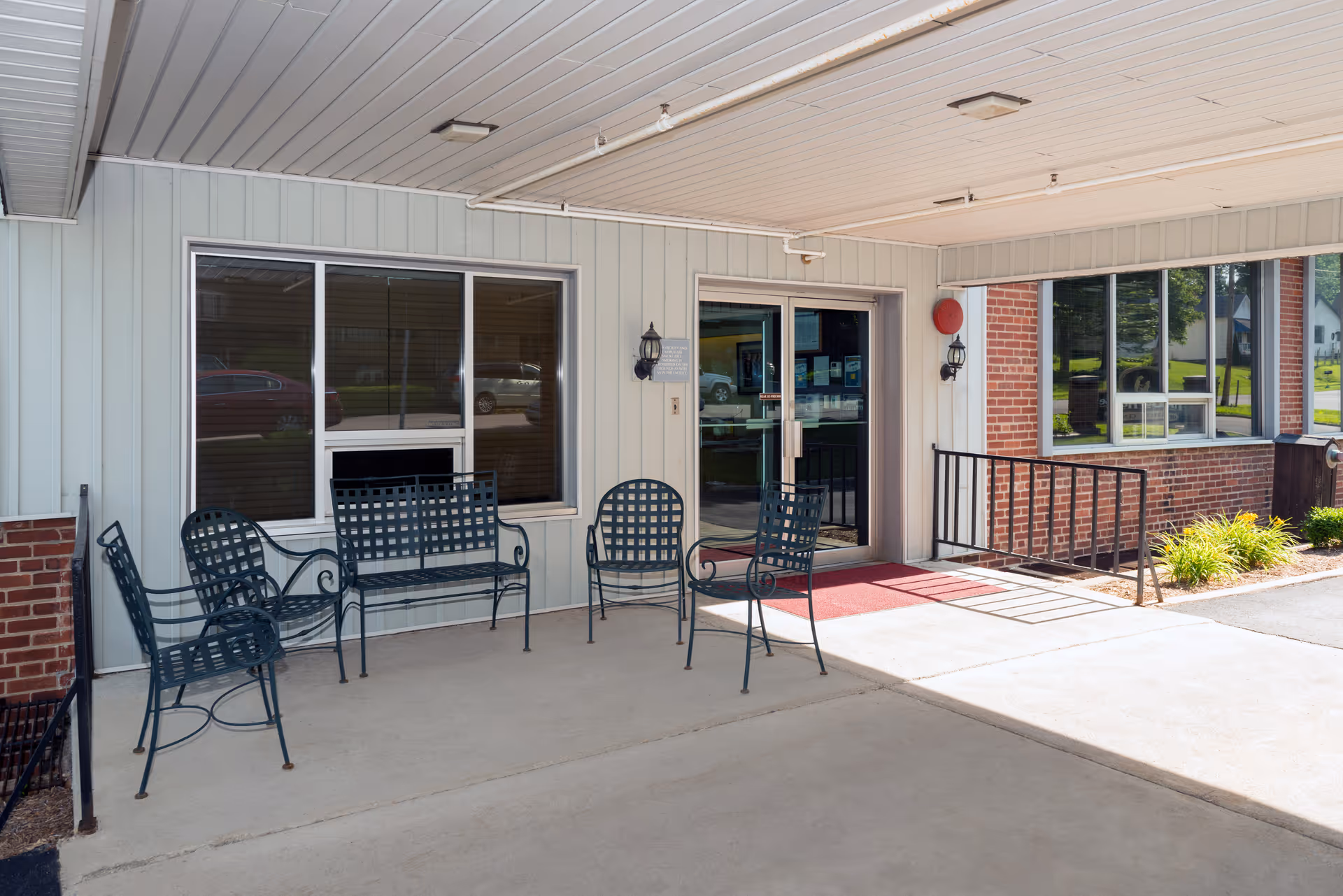 Covered entrance area of The Lane House with metal chairs and a bench arranged on the concrete floor. The building exterior features light-colored siding and brick walls, with large windows and a glass door leading inside. There is a red mat in front of the door and outdoor lighting fixtures on the walls.