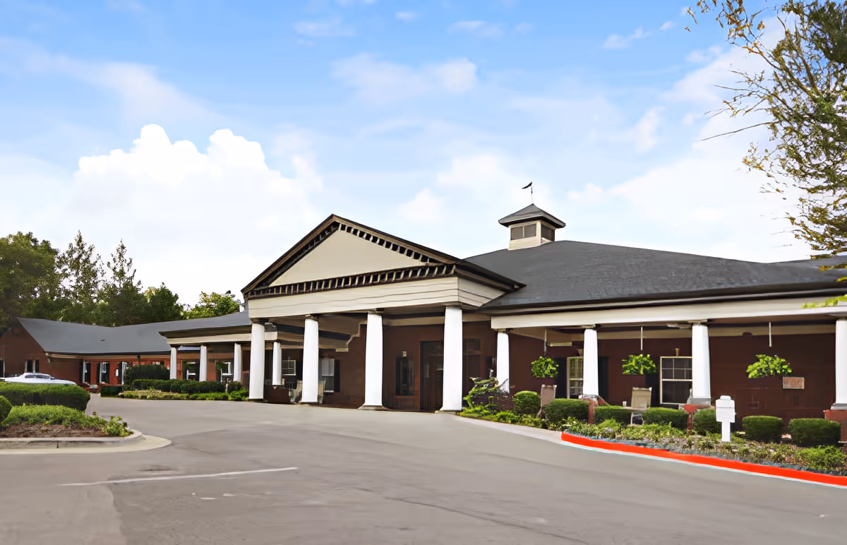 Entrance of a single-story brick senior living building with white columns, a covered porte-cochère, and landscaped grounds under a blue sky.