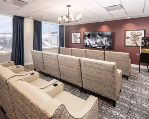 A cozy media room with two rows of beige and patterned reclining chairs facing a wall-mounted flat-screen TV playing a black and white movie. The room has a maroon accent wall with framed vintage film posters and a popcorn machine on the right side. Large windows with dark curtains allow natural light to enter the space.