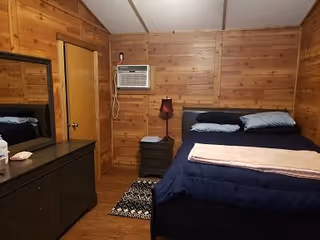 Wood-paneled bedroom with a bed dressed in blue bedding, a dresser with mirror, nightstand and a window air conditioner.