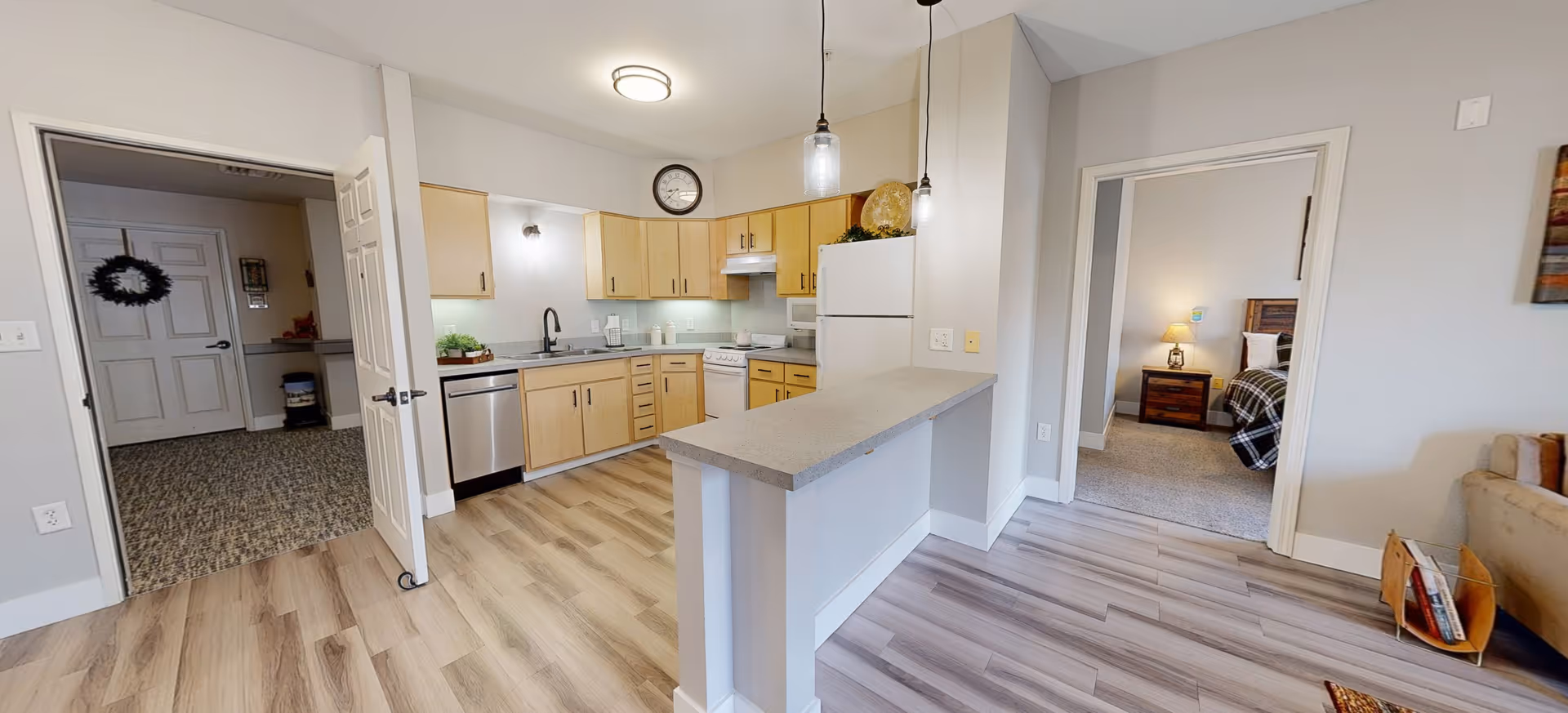 Interior view of a senior living facility apartment showing a kitchen with light wood cabinets, a white refrigerator, dishwasher, stove, and a countertop with two hanging pendant lights. To the right, there is a doorway leading to a bedroom with a wooden nightstand and lamp. To the left, there is an open door leading to a hallway with a carpeted floor and a door decorated with a wreath.