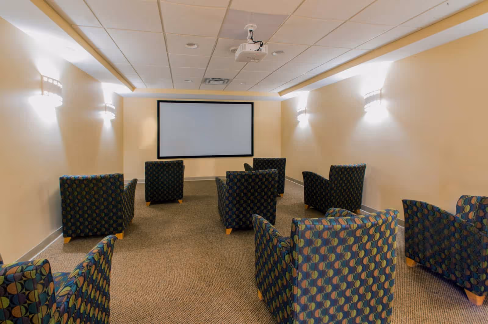 A small theater room with eight patterned armchairs arranged in rows facing a large blank projection screen on a beige wall. The room has a carpeted floor, cream-colored walls, ceiling lights, and a ceiling-mounted projector.