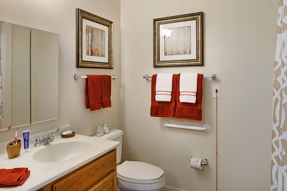 A clean bathroom with a white sink and wooden cabinet, a toilet, and red and white towels hanging on towel racks. There are two framed pictures on the wall above the towels, a mirror above the sink, and a patterned shower curtain visible on the right side.