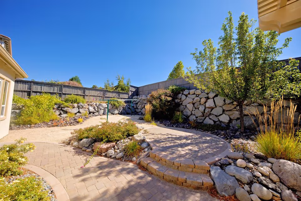 Sunny outdoor courtyard with stone wall, trees, and curved brick walkway.