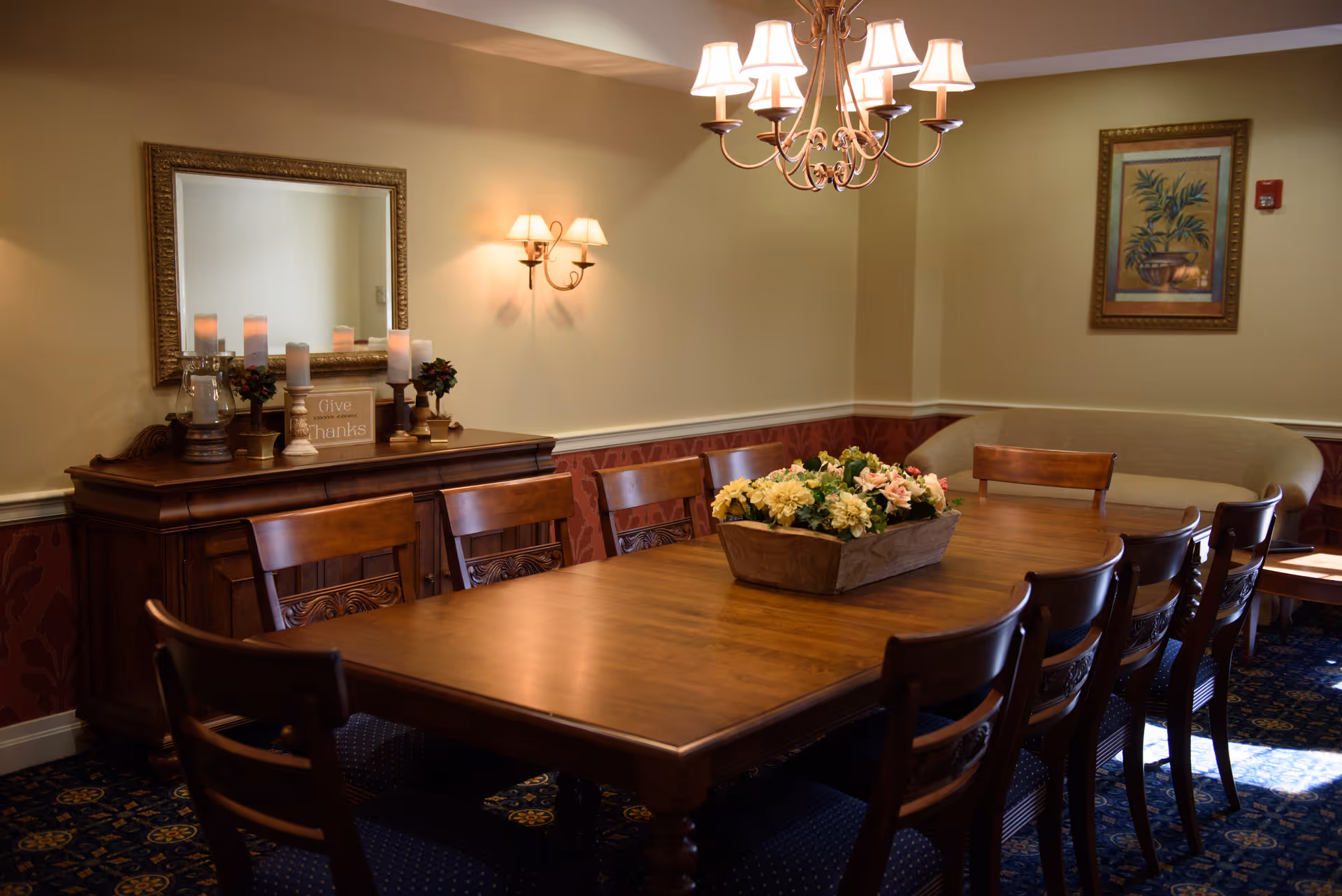 A warmly lit dining room with a long wooden table surrounded by wooden chairs. A floral centerpiece decorates the middle of the table. On the left side, there is a wooden sideboard with candles, small plants, and a sign that reads 'Give Thanks' beneath a large framed mirror. The walls are painted beige with a red patterned lower section, and there is a framed painting of a plant on the right wall. A chandelier with multiple lampshades hangs above the table.