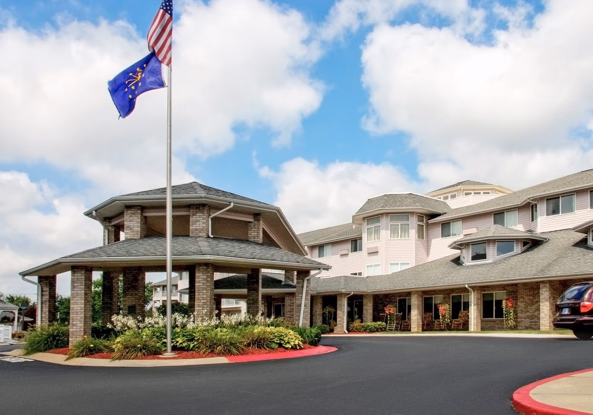 Exterior view of a senior living facility named Edgewood Downs with a circular driveway, a covered entrance supported by brick columns, landscaped greenery, and two flags on a flagpole against a partly cloudy blue sky.