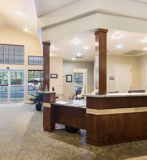 Reception area inside Meadow Creek Village Assisted Living with a wooden front desk, office chairs, and large windows letting in natural light. The space has beige walls and carpeted floors, with a glass entrance door visible in the background.