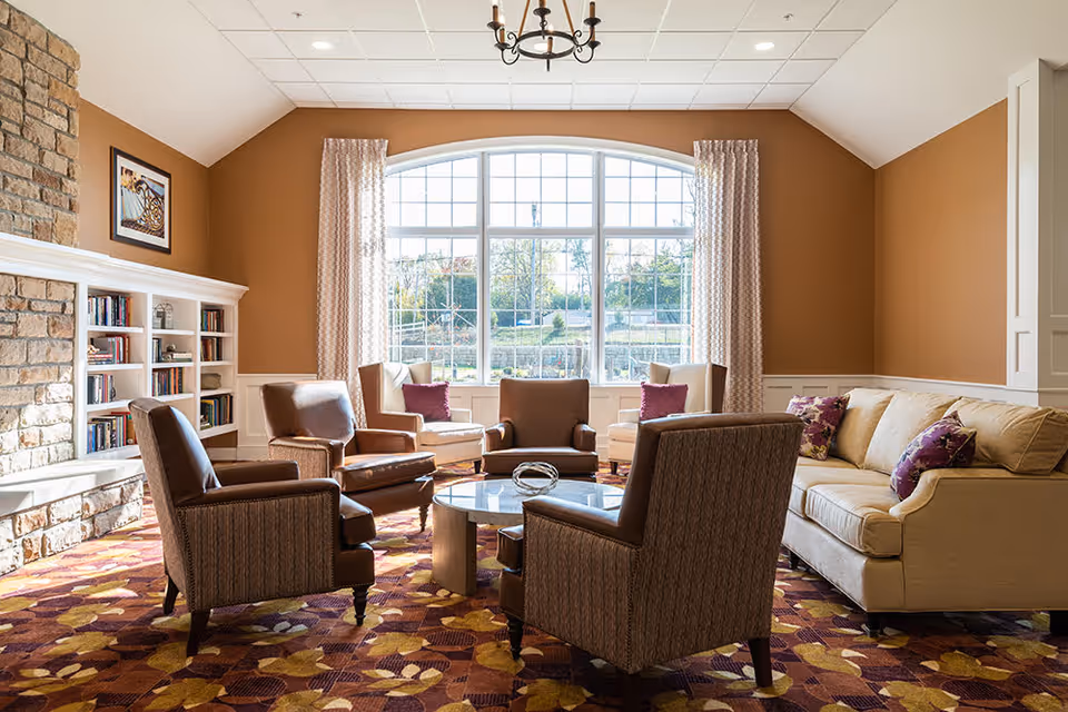 A cozy living room with a large arched window letting in natural light. The room features a beige sofa with purple floral pillows, four armchairs arranged around a round glass coffee table, a stone fireplace on the left, built-in white bookshelves, and warm brown walls with patterned curtains.