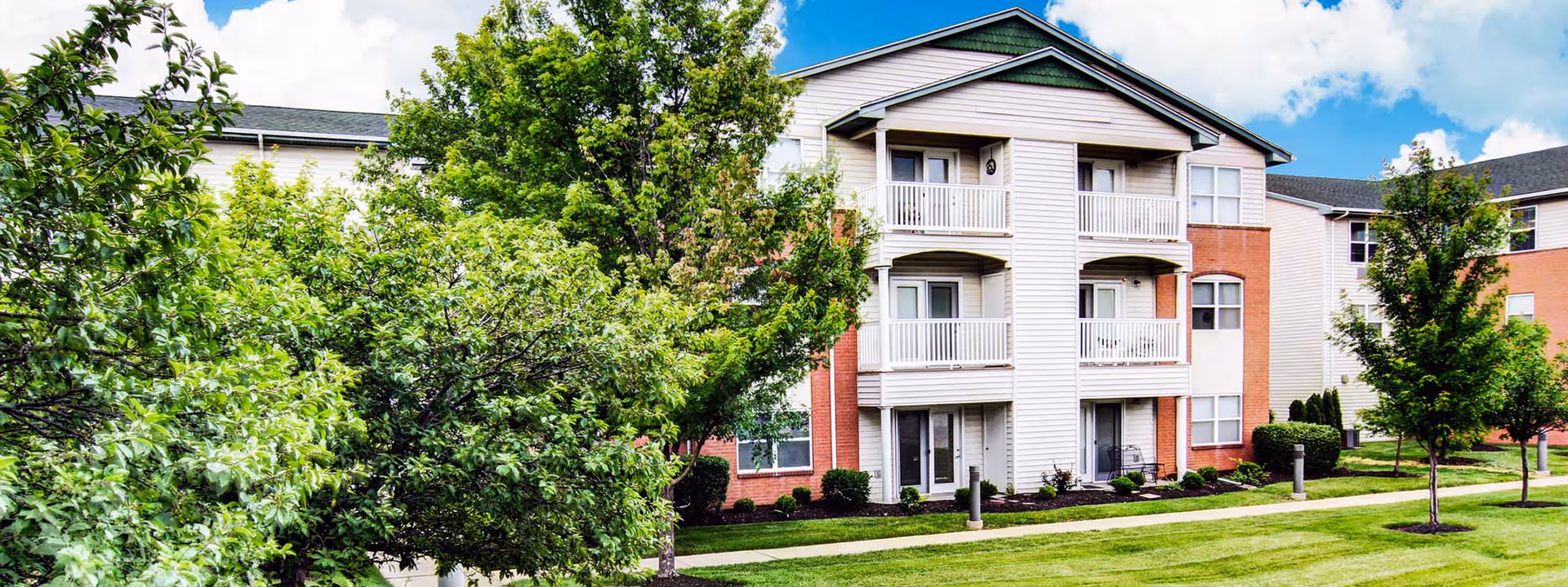 Three-story senior living building with balconies, landscaped lawn, trees, and walkways under a partly cloudy sky.