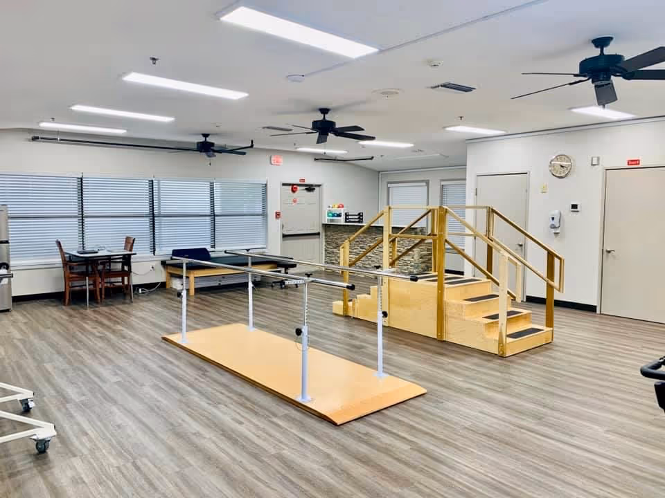 A spacious rehabilitation room with wooden flooring, parallel bars for walking exercises, a wooden staircase with handrails for physical therapy, ceiling fans, and a small table with chairs near large windows with blinds.