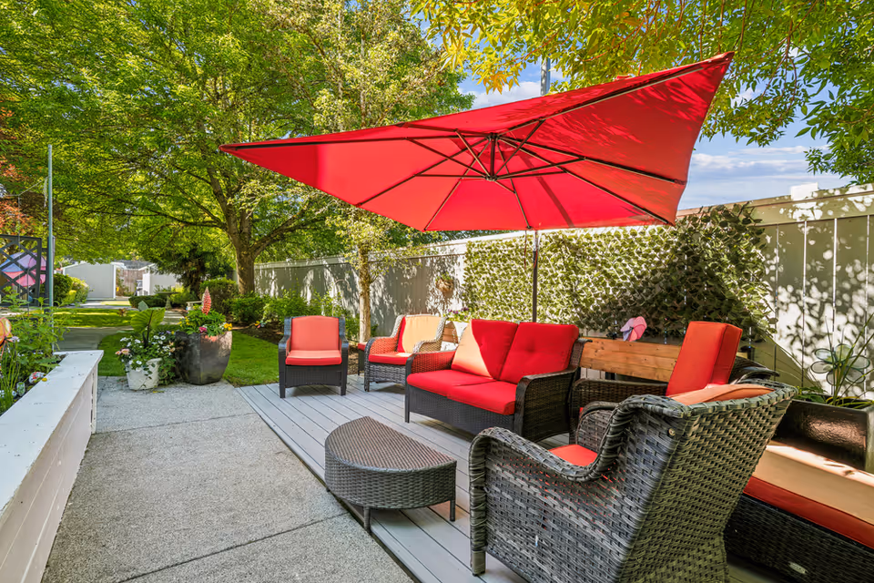 Outdoor patio area with wicker furniture including a sofa, chairs with red cushions, a small coffee table, and a large red umbrella providing shade. The patio is surrounded by greenery, trees, and a white fence with climbing plants.