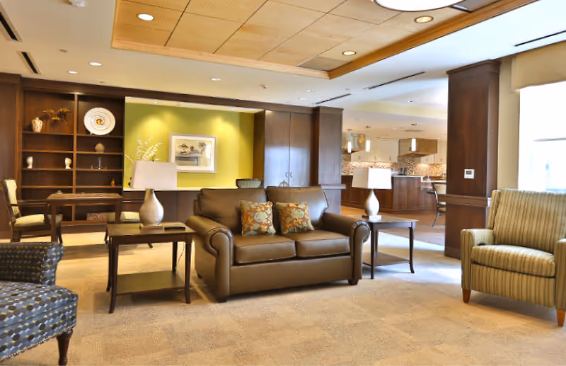Well-lit communal living area with a leather sofa, armchairs, side tables, bookshelves, and an open kitchen in the background.