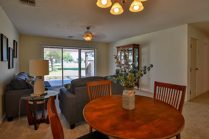 Interior view of a senior living facility's common area featuring a round wooden dining table with four chairs and a vase with flowers in the foreground, two gray sofas with patterned pillows, a side table with a lamp, a glass-front cabinet, and large sliding glass doors leading to an outdoor area with green grass and trees.