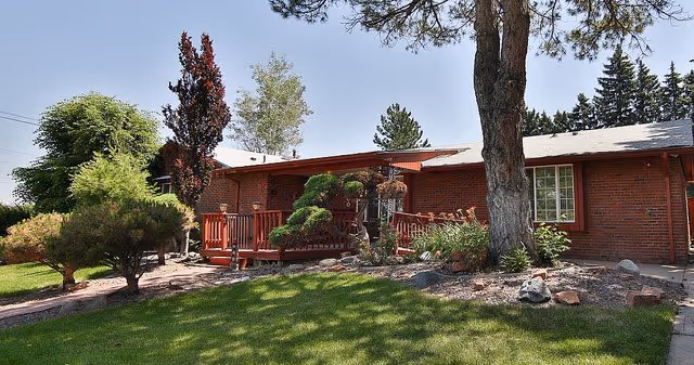 Single-story brick building with a red wooden porch surrounded by trees and shrubs, set against a clear blue sky.
