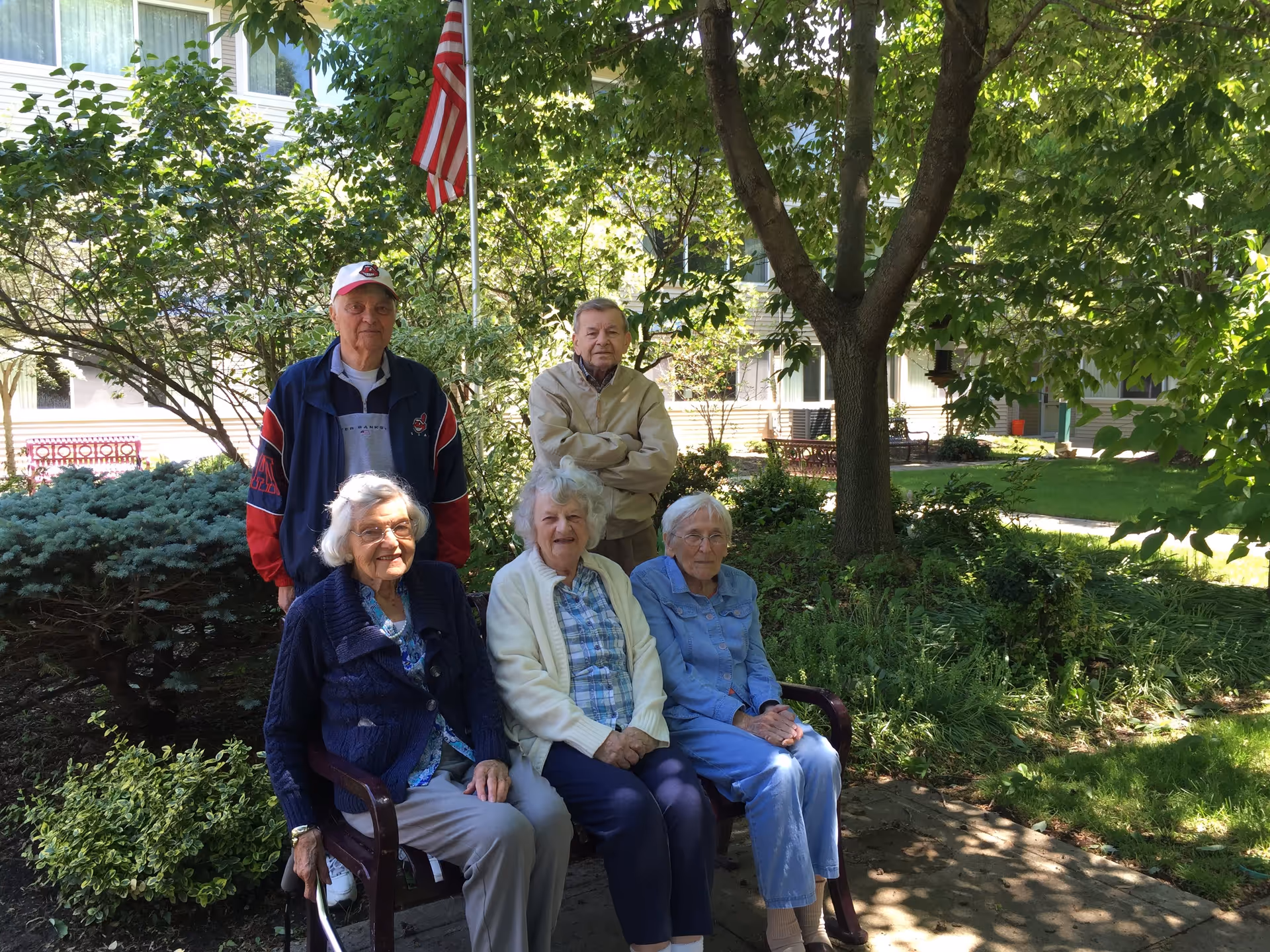 Five elderly people, three women sitting on a bench and two men standing behind them, are gathered outdoors in a garden area with trees, bushes, and an American flag in the background. The setting appears to be a peaceful courtyard of a retirement community.