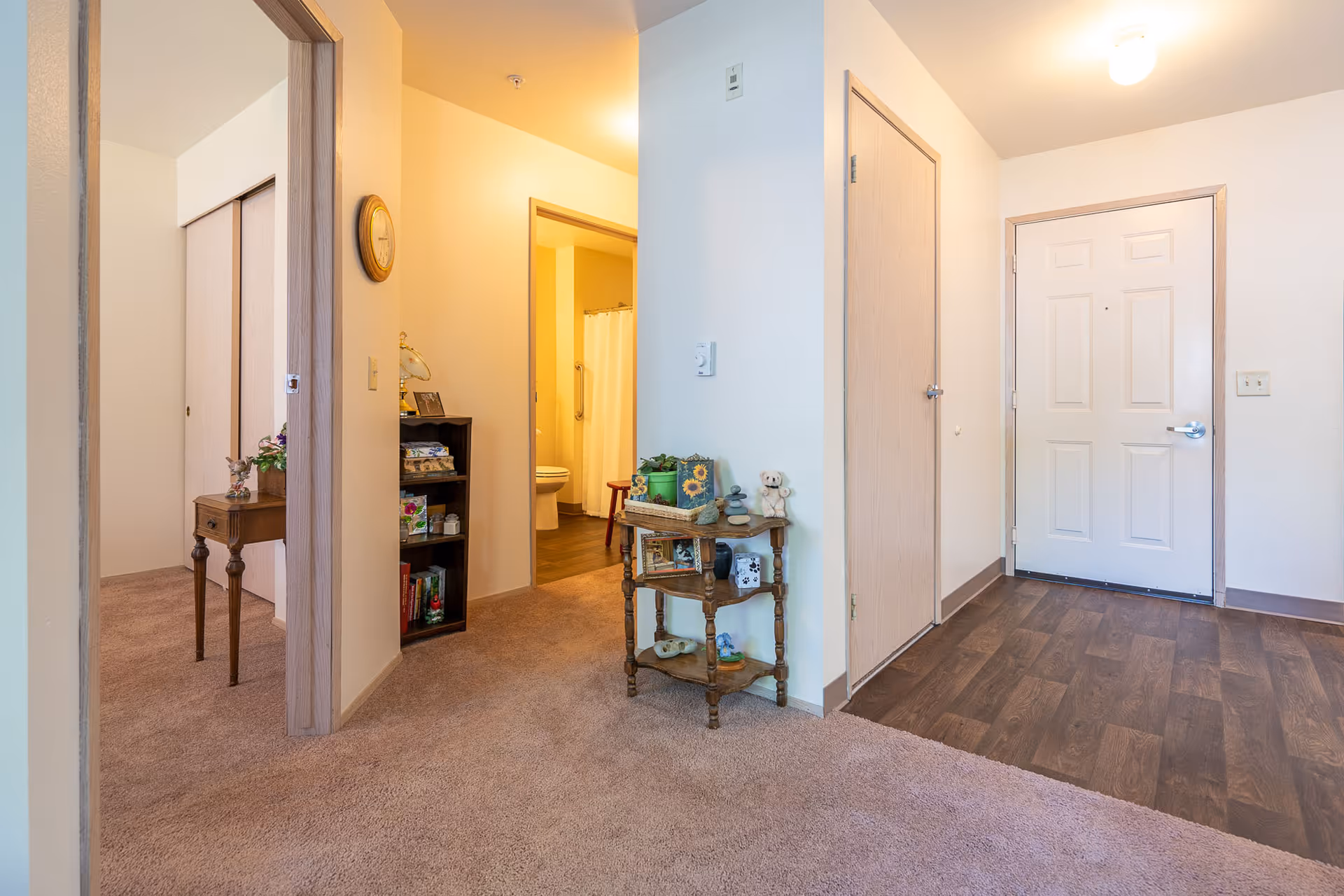 Interior view of a retirement facility showing a hallway with carpeted floor leading to a bathroom with a visible toilet and shower curtain. There is a small wooden table with decorative items and a bookshelf with books and other items. The entrance door with a wood-like floor is also visible.