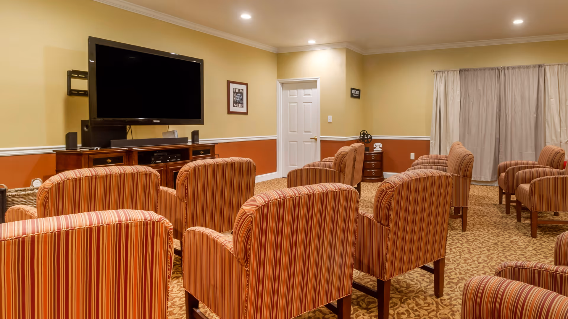 A cozy common room with multiple striped upholstered armchairs arranged in rows facing a large flat-screen TV mounted on a wooden cabinet. The room has beige walls with a white chair rail and a reddish-brown lower wall section, carpeted floor with a patterned design, a closed white door, framed wall art, and beige curtains covering a window.