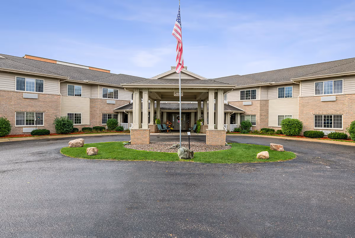 Front exterior view of a two-story senior living facility building with a covered entrance, an American flag on a flagpole in the center, and a circular driveway with landscaping including grass and rocks.