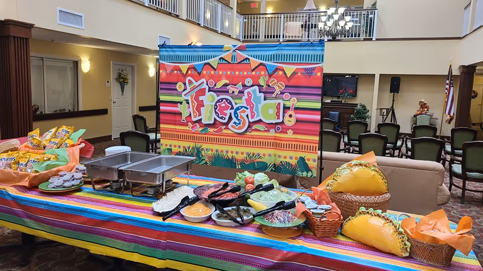 A festive buffet table set up in a senior living community common area with a colorful striped tablecloth and a bright 'Fiesta' banner. The table features various food items including chips, dips, and taco-shaped decorations. In the background, there are chairs, a TV, and an American flag in a spacious, well-lit room with a high ceiling and balcony.